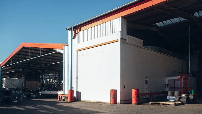 Zwei industrielle Lagergebäude mit roten Verkleidungen und Metalldächern stehen nebeneinander unter einem klaren blauen Himmel. Der Bereich zwischen ihnen ist gepflastert, und in der Nähe der Gebäude sind verschiedene Geräte und Paletten zu sehen.