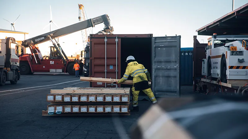 Ein Arbeiter in einem gelben Sicherheitsanzug und mit Helm bewegt einen Holzbalken in der Nähe von gestapelten Paletten in einem belebten Verschiffungshafen, mit Frachtcontainern, Kränen und Fahrzeugen im Hintergrund.