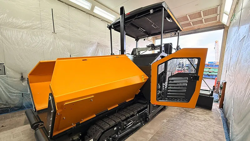 A large yellow and black construction vehicle, likely an asphalt paver, is parked in a warehouse with its driver's door open. The vehicle has tank-like tracks and is surrounded by white plastic tarps. 