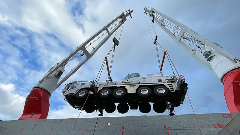 Ein großer mobiler Kranwagen wird von zwei Schwerlast-Schiffskränen hoch über ein Schiffsdeck gehoben, während der Himmel teilweise bewölkt ist.