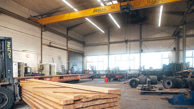 A spacious industrial hall with stacked wooden boards in the foreground, a forklift, tools on the walls, machine parts, and a yellow overhead crane labeled DEMAG 5t. Bright windows illuminate the room.