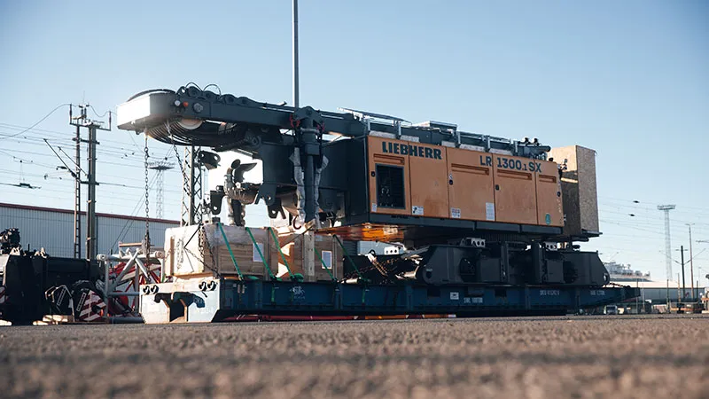 Eine große gelbe Liebherr-Krankomponente LR 1300.1 SX ist auf einem Tieflader gesichert und bereit für den Transport auf einem Industriegelände mit Gebäuden, Stromleitungen und Bahngleisen im Hintergrund.