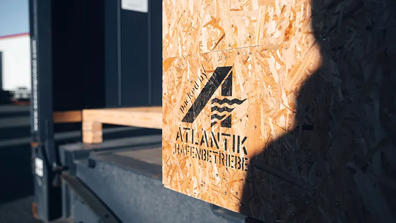 Nahaufnahme einer hölzernen Transportkiste mit dem Logo und dem Text ATLANTIK, die auf einer Laderampe steht und von der Sonne beschienen wird.