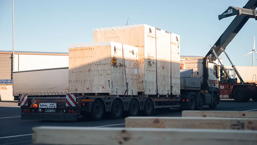 Ein Pritschenwagen ist in einem Industriegebiet geparkt und transportiert große Holzkisten. Im Hintergrund sind ein Kran und Schiffscontainer zu sehen, was auf einen Versand- oder Logistikbetrieb hindeutet.