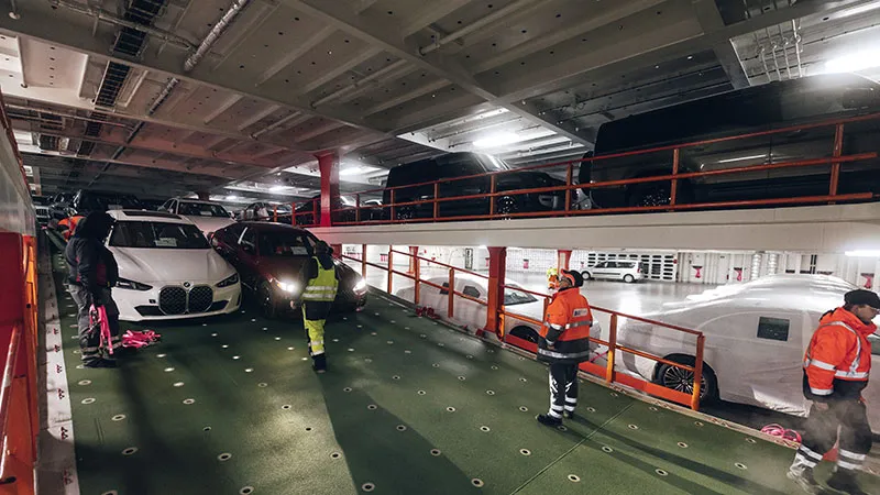Experience Workers in high-visibility clothing secure cars on a two-level vehicle deck inside a large ship, where various cars are parked closely together under artificial lighting.