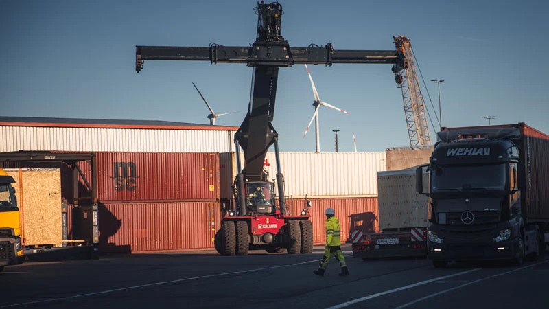 Know-How A worker in high-visibility clothing crosses a shipping yard where a large crane is moving a container. In the background, stacked shipping containers, a truck, and wind turbines are visible under clear skies.