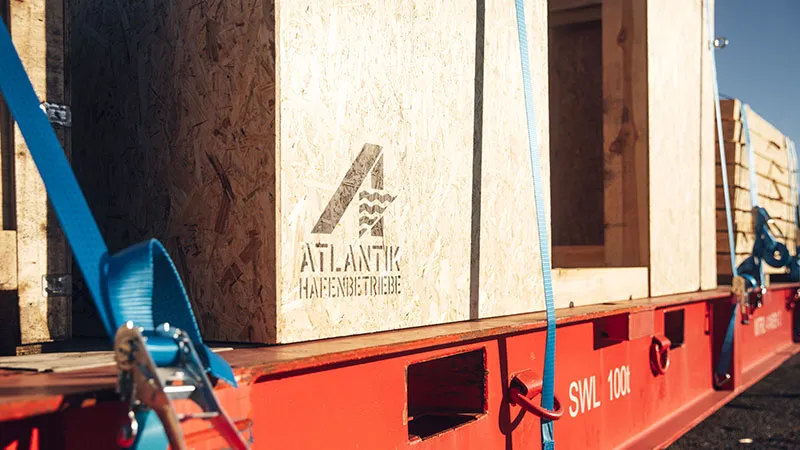 One-Stop Solutions Wooden crates bearing the Atlantik Hafenbetriebe logo are secured with blue straps on a red lowboy trailer for transport in bright sunshine.