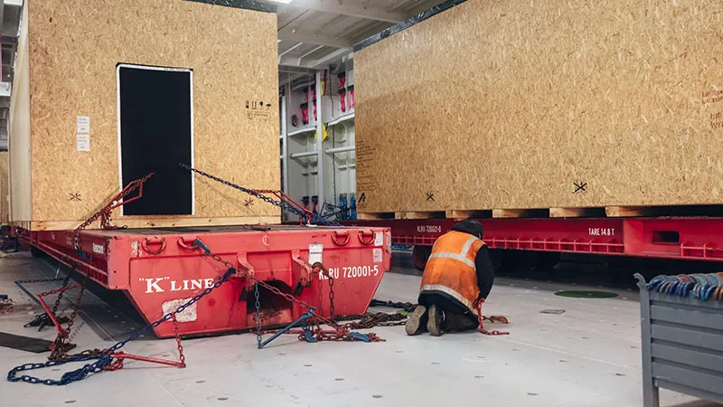 Ein Arbeiter in einer orangefarbenen Sicherheitsweste kniet auf dem Boden und befestigt große Holzkisten mit Ketten auf roten Flachbettcontainern in einer weitläufigen Industrieanlage.