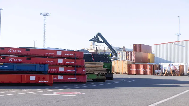 Technical services A shipping yard with stacked red and blue shipping containers, a crane lifting cargo, and various shipping containers in the background near a warehouse building.