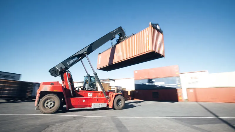 Terminal and CFS services A red forklift lifts a large shipping container in a shipping yard, with other stacked containers and a clear blue sky in the background.